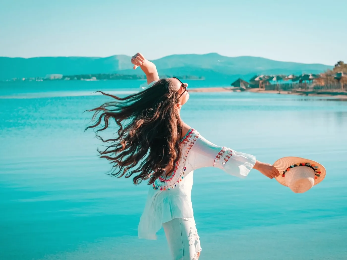 A woman with flowing hair stands on the beach, arms wide open, enjoying the sun and the sound of the waves.