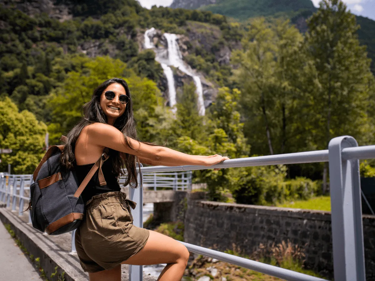 Une femme portant des lunettes de soleil et un sac à dos est assise sur une rambarde près d'une cascade.