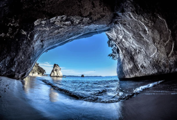 Vue depuis l'intérieur d'une grotte sur la plage, avec des vagues et des rochers en arrière-plan, créant une atmosphère paisible.