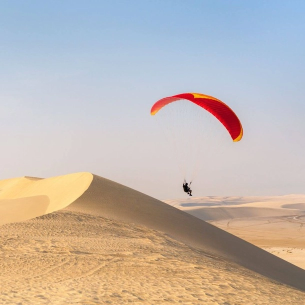 Une personne en parapente survole des dunes de sable, offrant une vue spectaculaire sur le paysage désertique.