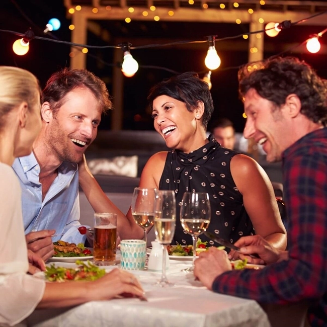 A group of people sitting around a table, sharing drinks and a variety of dishes in a convivial atmosphere.