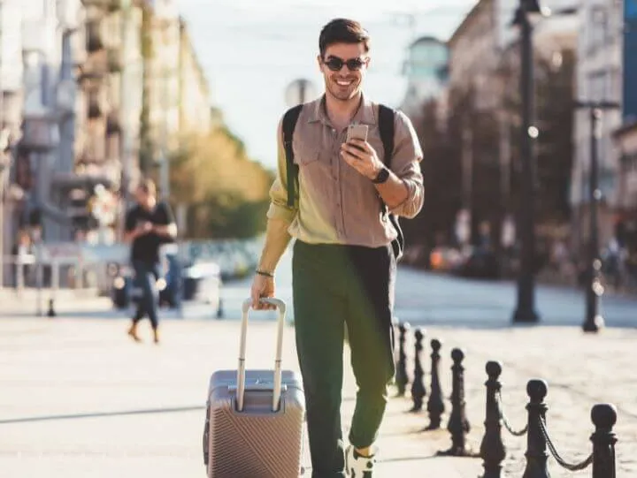 Un homme marchant dans la rue, tirant une valise derrière lui, sous un ciel dégagé.