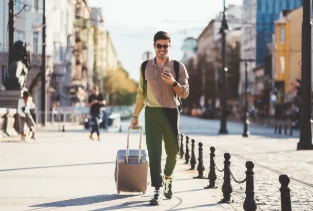 A man walking down the street, pulling a suitcase behind him, under a clear sky.