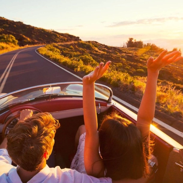 Un couple souriant conduit une voiture décapotable, les mains levées, profitant d'une belle journée ensoleillée.