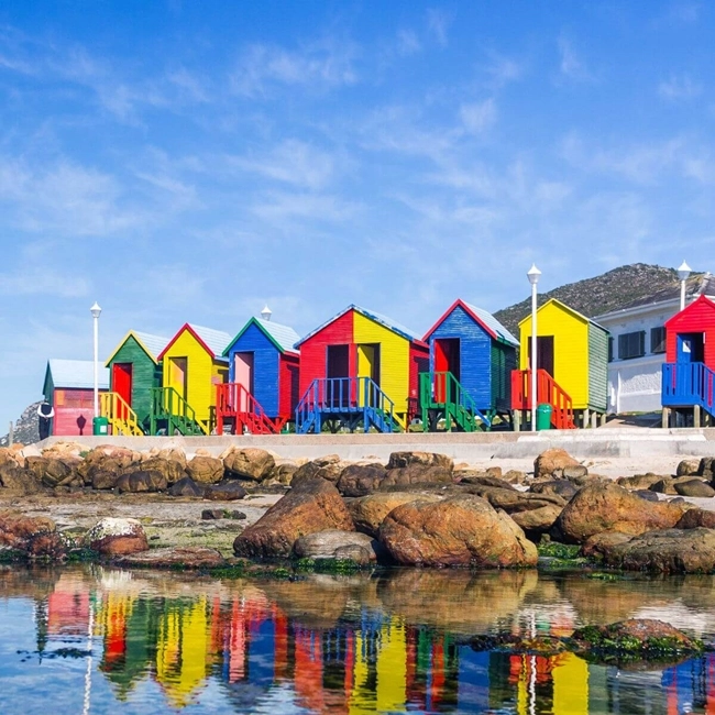 Colorful beach huts line the shore of a body of water, creating a vibrant, welcoming landscape.