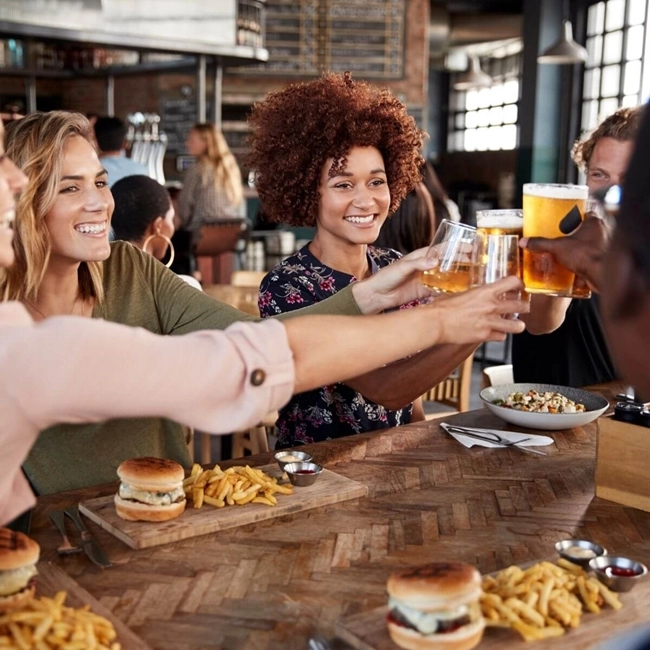 A group of people raise a glass of beer in celebration around a table set with tasty dishes.