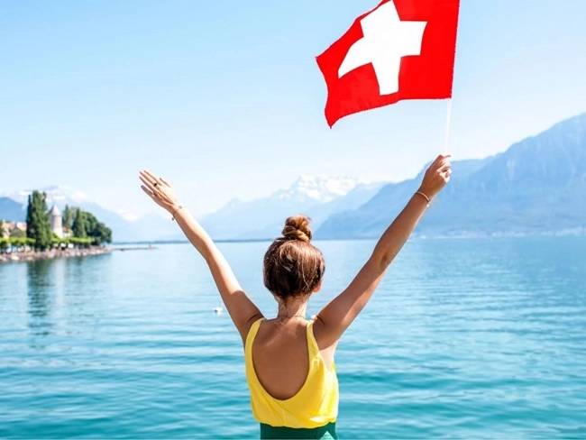 A woman holding a Swiss flag in front of a peaceful lake, symbolizing Switzerland's natural beauty.