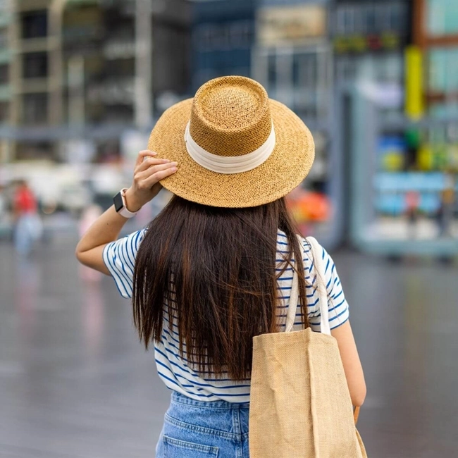 Rear view of a young woman wearing a straw hat and striped shirt, strolling through the city.