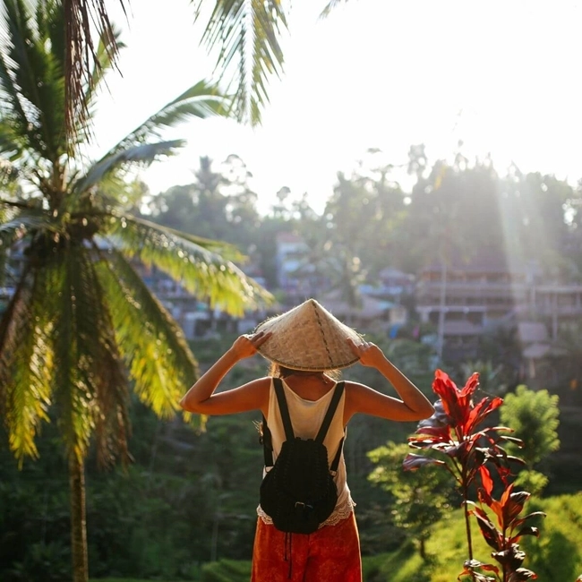 A woman wearing a hat and shorts stands on a hill, admiring a picturesque village below.